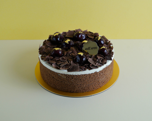 A black forest cake with layers of rich chocolate sponge, whipped cream, and cherries. The cake is topped with dark chocolate shavings and garnished with fresh cherries.