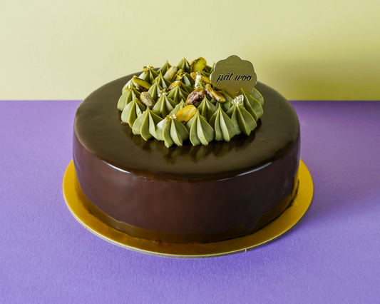 Cake with dark chocolate glaze garnished with pistachios and pistachio whipped cream. The cake is layered with light pistachio sponge and creamy Sicilian pistachio filing.