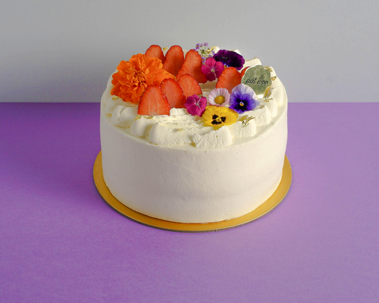 An elegant layered cake decorated with fresh strawberries and edible flowers, coated with Madagascan vanilla bean cream which are known for their rich, smooth texture and distinctively sweet and floral flavour. The cake also features fluffy vanilla cake layers and fresh strawberries, reminiscent of the classic strawberry shortcake dessert. 